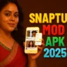 A woman with long, dark, wavy hair, adorned with white flowers on the right side, wearing a red and gold sari and large earrings, stands slightly to the left, looking directly at the viewer and smiling. She holds a yellow smartphone in her left hand, displaying a video streaming interface with various video thumbnails and an overlay menu. To the right, in large, colorful, sans-serif letters, are the words "SNAPTUBE" in yellow, "MOD" in red, "APK" in green, and "2025" in teal. The background is a solid, dark reddish-brown color.