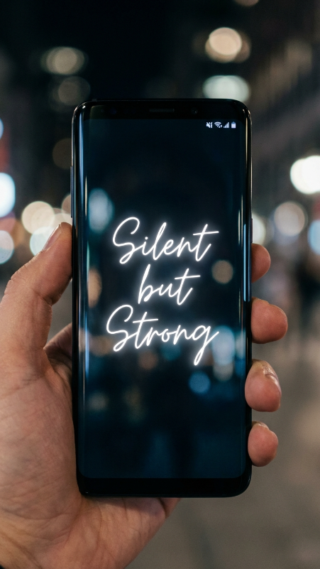 Ultra-realistic smartphone held vertically in a hand, modern black Android lock screen visible with elegant glowing handwritten text in the center reading “Silent but Strong”, soft dark wallpaper background with blurred city lights at night, realistic screen reflections, premium mobile frame, minimal icons at top, cinematic lighting, highly realistic phone mockup, no extra text outside the screen.